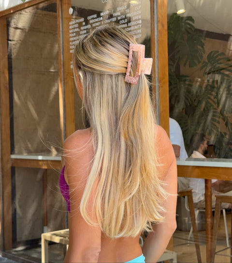 A woman viewed from behind wearing bright turquoise leggings and a pink hair clip, standing in front of a cafe with large glass windows and wooden frames.